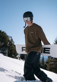 Snowboardeur vêtu d'un sweat-shirt marron et d'un pantalon noir, portant un casque noir avec des lunettes de protection miroitées, transportant un snowboard blanc avec des lettres noires.
