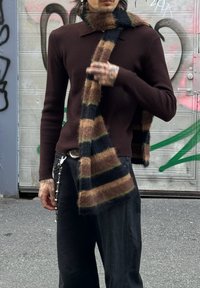 Pull en maille marron avec col, associé à une écharpe rayée marron, noire et verte. Comporte un pantalon noir avec un accessoire chaîne.