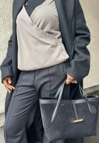 Άτομο που φοράει μαύρο παλτό, τοπ τύπου wrap σε αποχρώσεις του ταυπ, μαύρο παντελόνι, κρατώντας μια μαύρη τσάντα tote από σουέτ και δέρμα με χρυσές λεπτομέρειες.