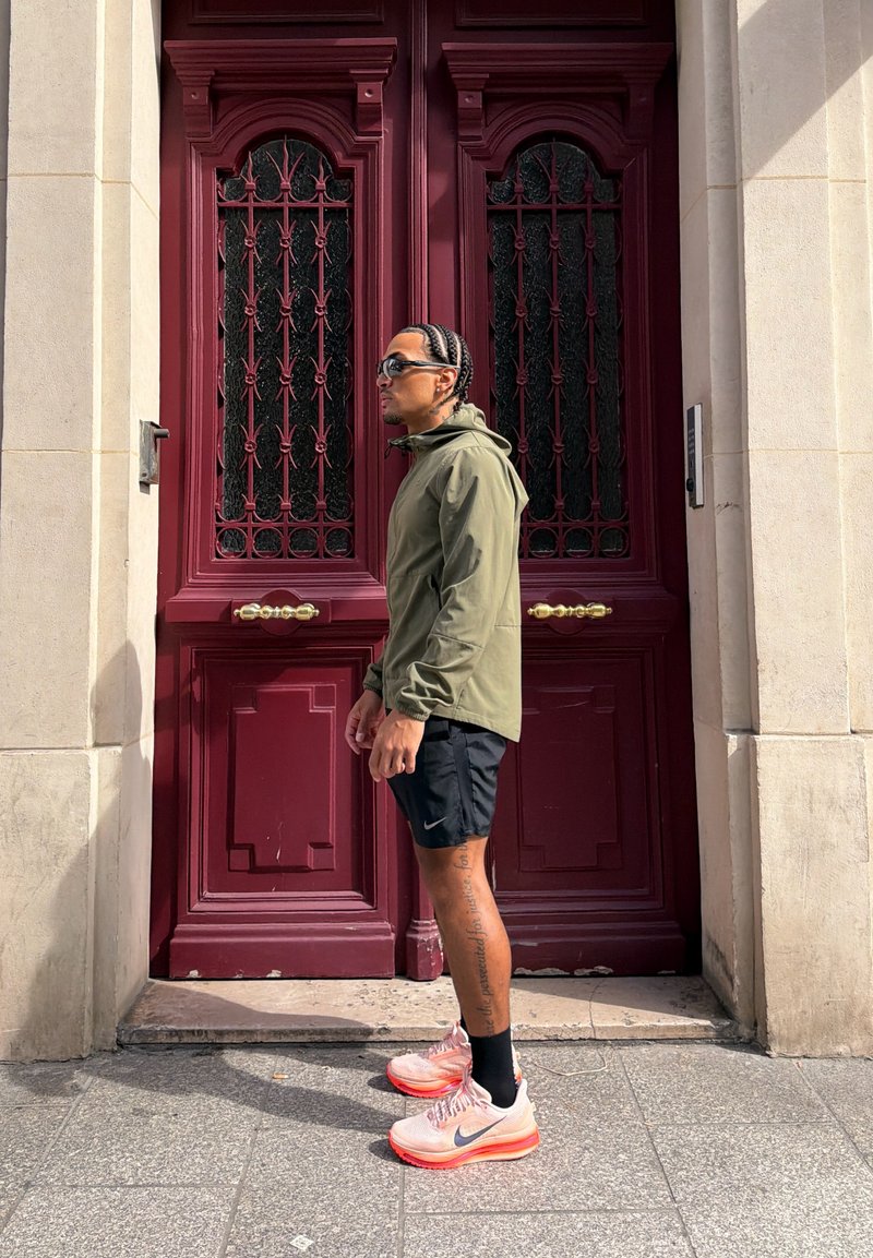 Homme aux cheveux tressés portant des lunettes de soleil, une veste verte, un short noir et des baskets roses, debout de côté devant une grande porte rouge.