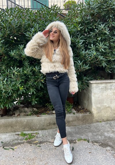 Chaqueta corta de piel sintética en crema con capucha, combinada con jeans skinny negros y zapatillas blancas. Follaje verde de fondo.
