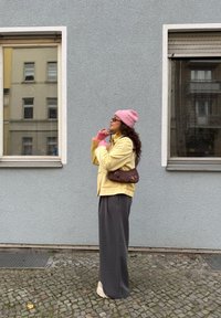 Persona che indossa una giacca gialla, un berretto rosa e guanti rosa, in piedi accanto a un muro grigio e grandi finestre, con pantaloni grigi e scarpe beige.