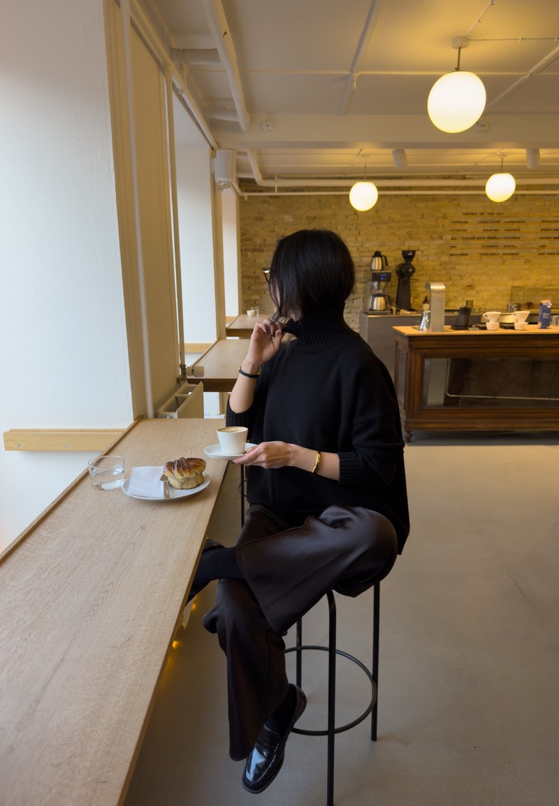 Pull noir à col roulé, chaussures noires vernies et pantalon en satin foncé. Table avec un verre d'eau, une tasse de café et une pâtisserie sur une assiette.