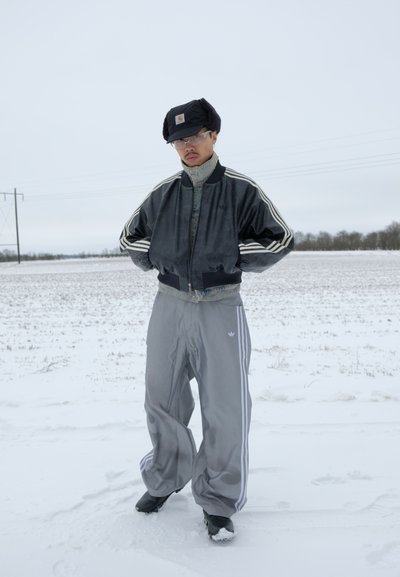 Hombre de pie con las manos en los bolsillos de la chaqueta en un campo nevado, vistiendo una chaqueta bomber negra, pantalones deportivos grises, gorro de invierno negro y gafas.