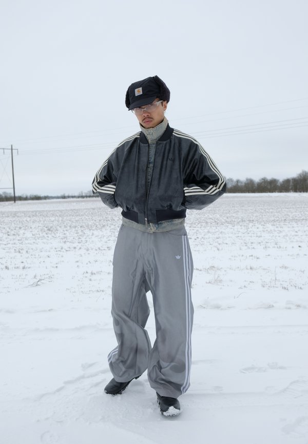 Hombre de pie con las manos en los bolsillos de la chaqueta en un campo nevado, vistiendo una chaqueta bomber negra, pantalones deportivos grises, gorro de invierno negro y gafas.