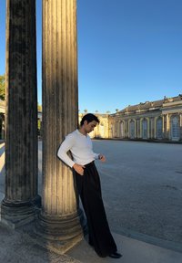 Mann in weißem Langarmshirt und schwarzen weiten Hosen, der sich gegen eine große Steinsäule in einem sonnenbeschienenen historischen Innenhof unter klarem blauen Himmel lehnt.