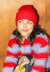 Jeune femme portant un bonnet en tricot rouge et un pull rayé rouge et lavande, assise contre un fond en bois, regardant sur le côté avec un léger sourire.