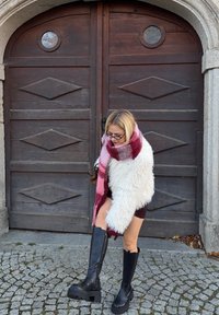 Sorte knæhøje plateau-støvler med en glat overflade, kombineret med en hvid fluffy sweater og et lyserødt ternet tørklæde. Stengårdsbelagt brostensbelægning.