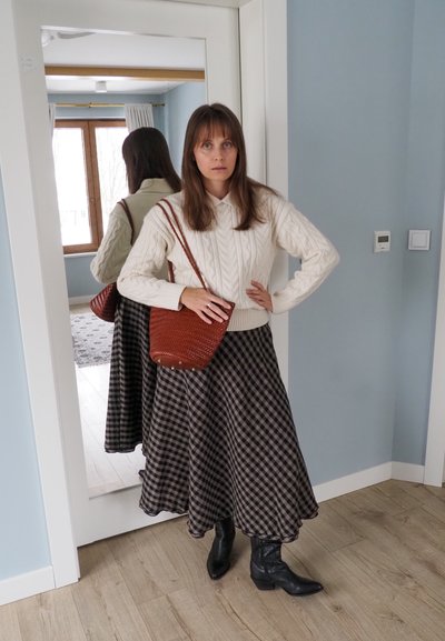 Mujer con suéter blanco de punto trenzado, falda midi de cuadros, botas negras y bolso marrón tejido al hombro, de pie junto a un espejo de cuerpo entero en una habitación azul claro.