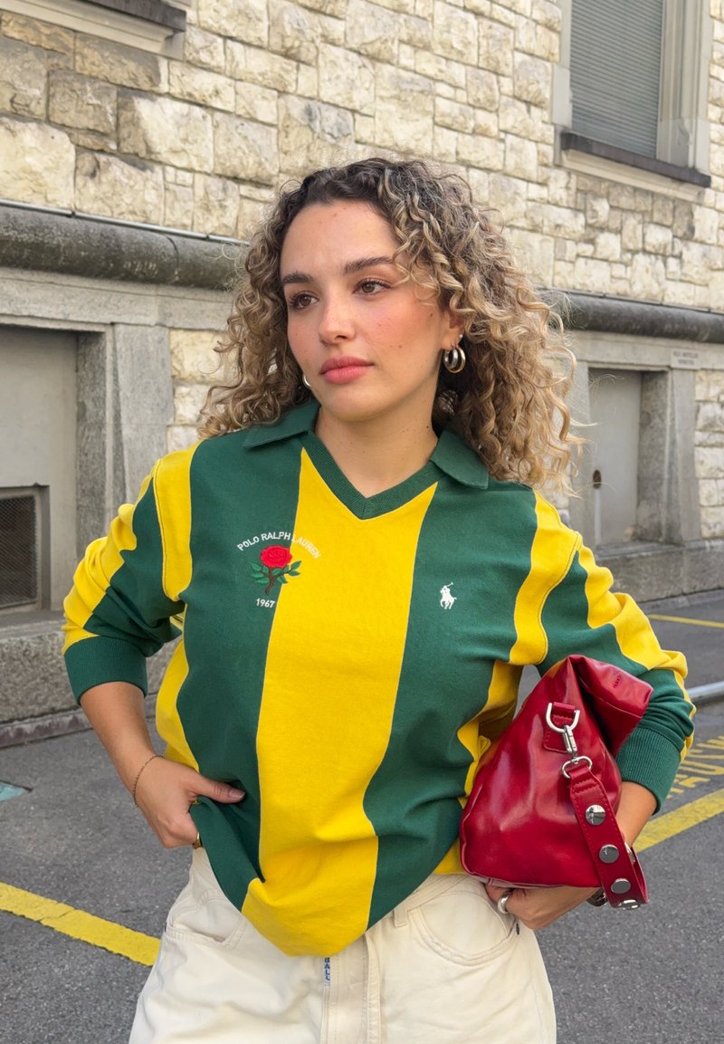 Junge Frau mit lockigem Haar, die einen grün-gelb gestreiften Polo Ralph Lauren Pullover trägt, hält eine rote Handtasche und steht im Freien.