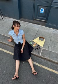 Camicetta azzurra a maniche corte, gonna nera fino al ginocchio, borsa gialla, drink verde, macchina fotografica, occhiali da sole e uno spuntino sul marciapiede.