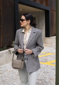 Šedý kostkovaný blazer přes béžovou blůzu, spárovaný se světle modrými džíny a texturovanou béžovou kabelkou. Černé sluneční brýle a zlaté náušnice.