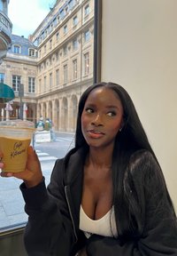 Kall kaffe i en klar kopp märkt "Café Kitumi", hålls av en person i en svart hette. Ljusa omgivningar med byggnader synliga genom ett fönster.