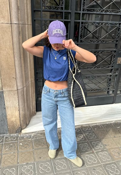 Camiseta corta azul, vaqueros de pierna ancha celestes, zapatos verde oliva, bolsa negra tejida con acentos crema, gorra morada con letras blancas.