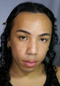 Persona con capelli neri ricci di media lunghezza, che indossa orecchini a cerchio dorati e un piercing al setto dorato, pelle chiara con lentiggini visibili.