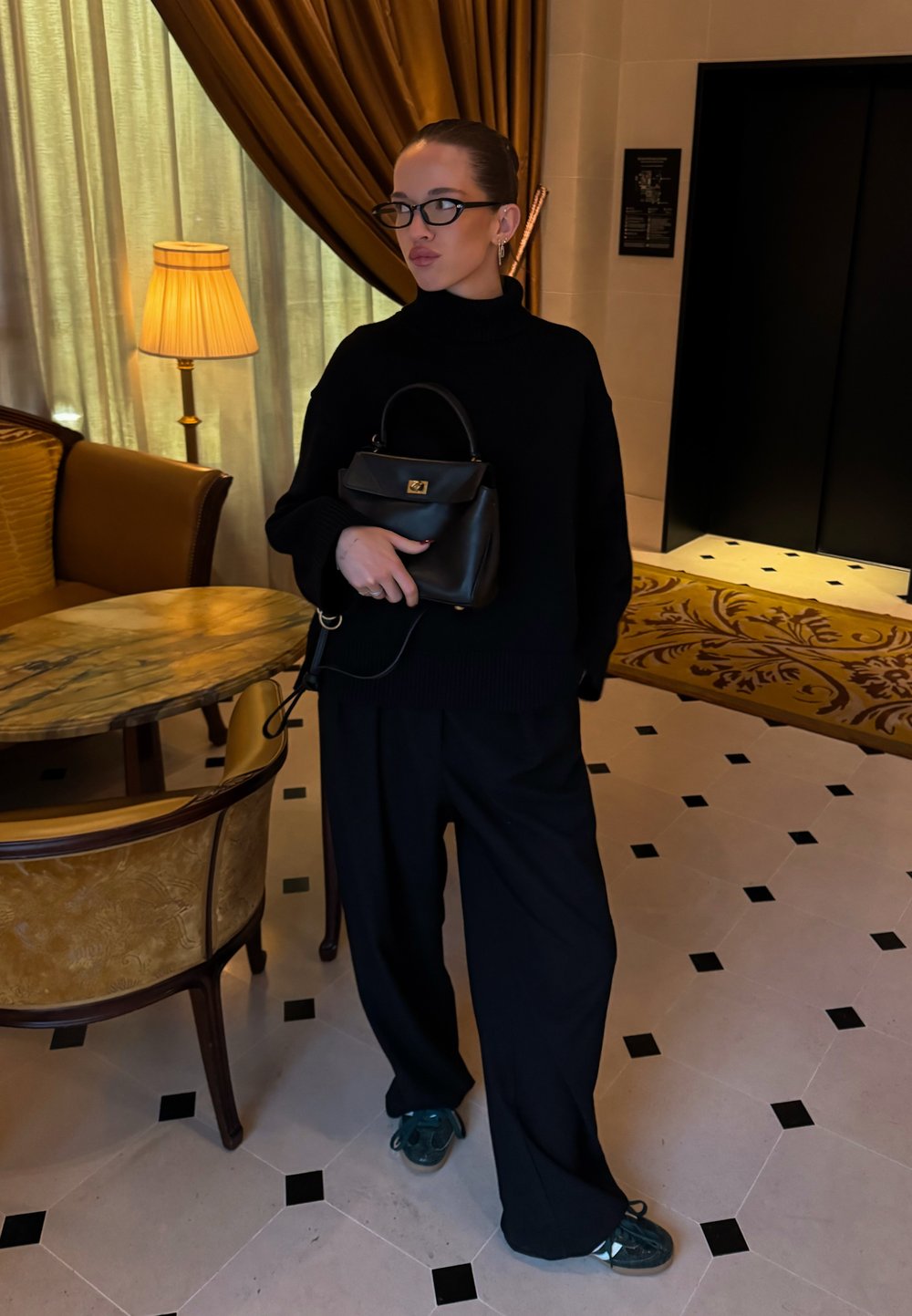 Pull noir à col roulé, pantalon noir ample et sac à main noir avec un détail doré. Le décor comprend un mobilier élégant et des carreaux de sol à motifs.
