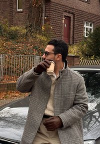 Grå og svart rutete jakke, kremfarget genser under, brune skinnhansker, holder en kaffekopp, står ved siden av en bil med en murvegg i bakgrunnen.