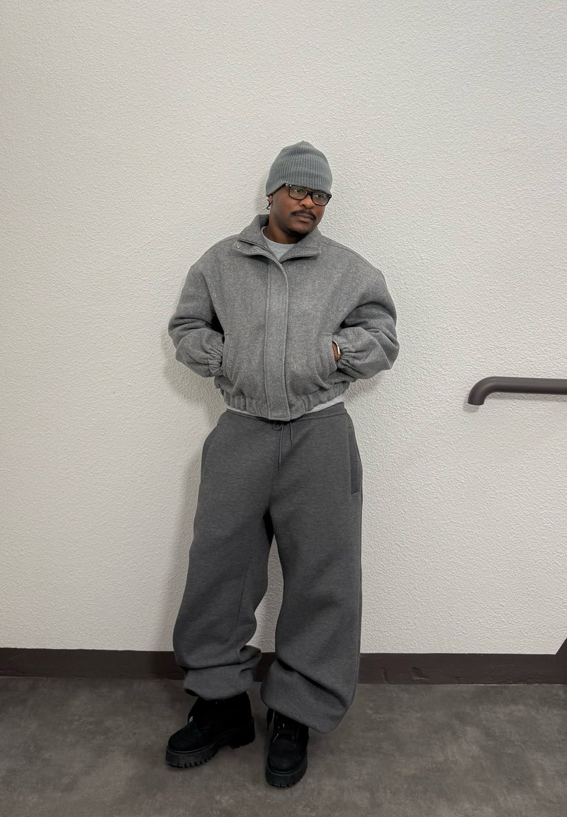 Homme portant un bonnet gris, des lunettes, une veste zippée grise, un pantalon gris ample et des bottes noires, debout les mains dans les poches contre un mur texturé.