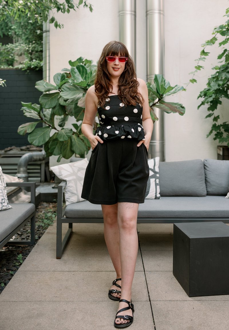 Femme portant un haut noir à pois, une jupe noire et des lunettes de soleil rouges, debout les mains dans les poches sur une terrasse extérieure avec des canapés gris et des plantes vertes.