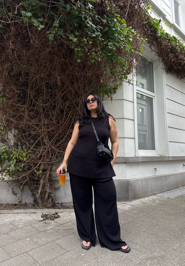 Γυναίκα με μαύρα ρούχα και γυαλιά ηλίου κρατάει ένα ποτό και στέκεται σε πεζοδρόμιο πόλης κοντά σε έναν τοίχο καλυμμένο με ξερά και πράσινα αναρριχητικά φυτά.