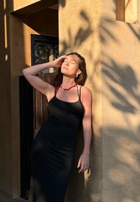 Femme en robe noire avec un collier rouge, les yeux fermés, la main touchant ses cheveux, appuyée contre un mur texturé baigné de lumière.