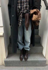 Personne debout sur des marches en pierre, portant un manteau foncé, une chemise à carreaux, un jean clair, des bottes marron, tenant un sac à main marron clair souple, et un pull marron noué à la taille.