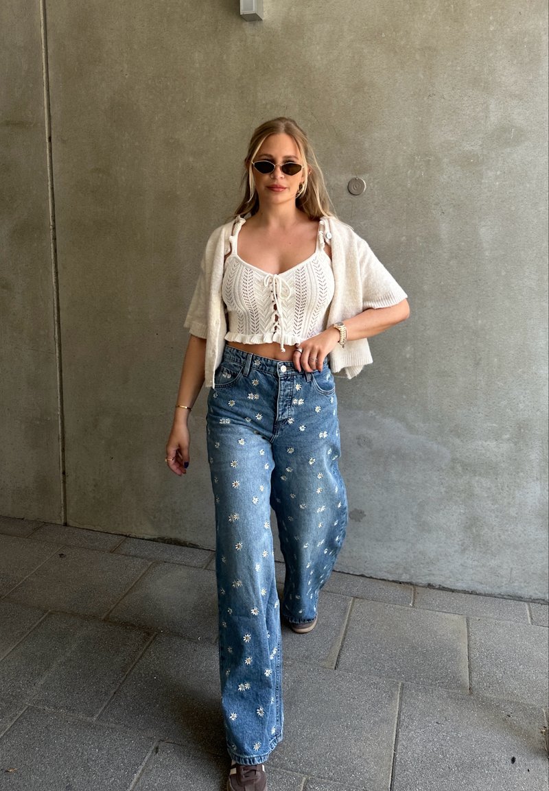 Cropped top bianco a maglia con allacciatura frontale, abbinato a jeans a gamba larga blu con ricami floreali. Scarpe marroni completano l'outfit.
