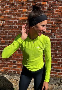 Maglietta sportiva a maniche lunghe, di un vivace verde lime, con tessuto testurizzato e logo, abbinata a leggins neri e una fascia nera contro un muro di mattoni.