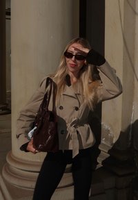 Trench coat beige chiaro con bottoni doppi e vita legata, abbinato a leggins neri e una borsa marrone scuro. Occhiali da sole neri.