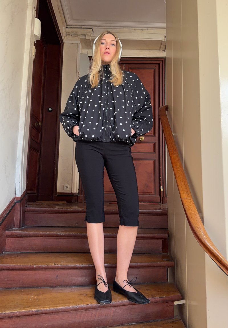 Jeune femme portant un casque blanc, une veste noire à pois, un pantalon noir court et des ballerines noires, debout sur des escaliers en bois à l'intérieur.