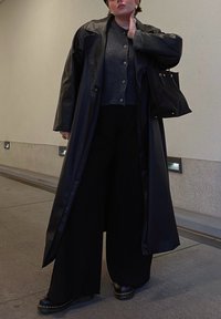 Μαύρο δερμάτινο παλτό μέχρι το γόνατο πάνω από γκρι πουλόβερ με κουμπιά και σκούρο φαρδύ παντελόνι. Μαύρα παπούτσια και τσάντα με υφή.