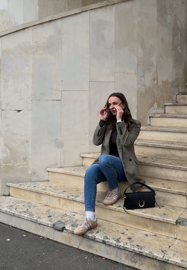 Rūtainā žakete, melna blūze, zilas džinses un bēšas sneakers. Sēž uz akmens pakāpieniem ar melnu somu. Dabas akmens sienas fons.