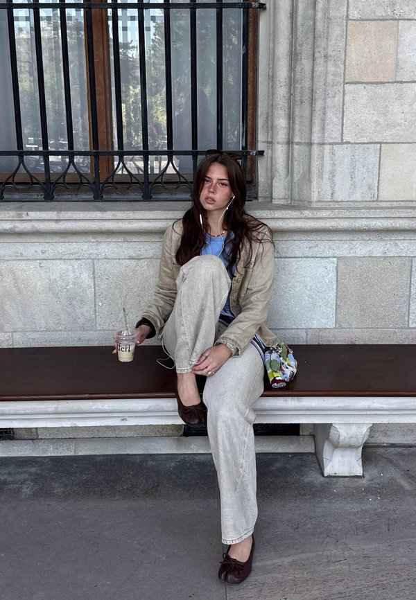 Jeune femme aux longs cheveux foncés portant une veste et un pantalon beige, assise sur un banc, tenant une boisson glacée et portant des écouteurs près d'un mur de pierre avec des barreaux de fenêtre.