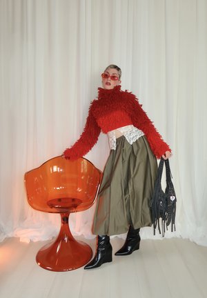 Mujer con suéter rojo texturizado, pantalones anchos color oliva, botas negras y gafas de sol rojas, apoyada en una silla translúcida naranja mientras sostiene una bolsa negra.