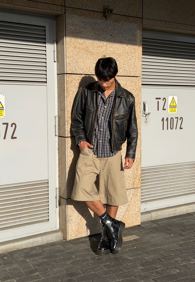 Jeune homme en veste en cuir noire, chemise à carreaux et shorts beige, appuyé contre un mur entre deux portes métalliques grises au soleil.