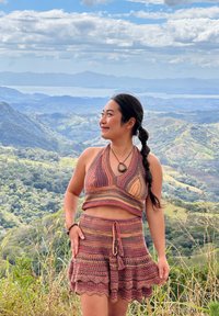 Femme portant un haut à bretelles et une jupe en crochet marron et orange, souriante, avec des montagnes, de la verdure et un ciel partiellement nuageux en arrière-plan.