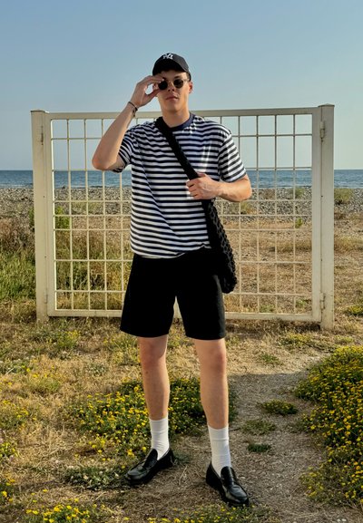 Camisa de rayas de manga corta, pantalones cortos negros, gafas de sol y un bolso negro de hombro. De pie cerca de una valla blanca de rejilla con un fondo de playa.