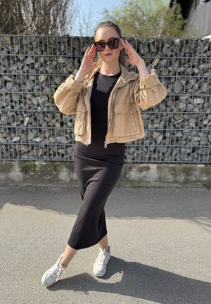 Chaqueta corta beige con cinturón sobre una blusa negra de manga larga y una falda midi negra, combinada con zapatillas blancas y gafas de sol oversized.