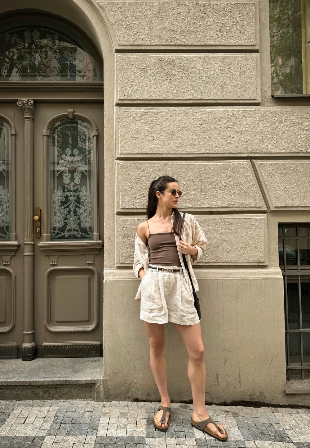 Light beige linen shorts, brown tank top, and a striped open shirt. Finished with circular sunglasses and brown flip-flops. Urban setting.