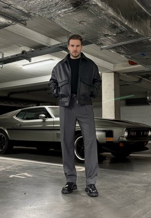 Chaqueta bomber de cuero negra, camisa negra, pantalones grises y zapatos negros. Clásico coche musculoso en el fondo sobre una superficie de aparcamiento de hormigón.