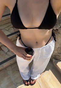 Fekete háromszög bikini felső, fehér laza nadrág, fekete papucs. Kéz egy elviteles kávéspoharat tart. Természetes kő háttér.