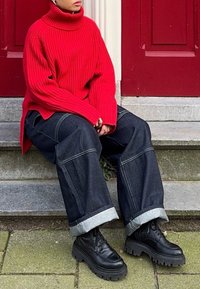 Maglione a collo alto rosso a costine, pantaloni larghi in denim scuro con cuciture a contrasto e risvolti, abbinati a stivali neri con lacci spessi.