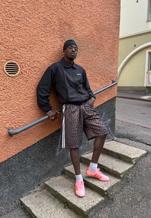 Chaqueta negra con cremallera, pantalones cortos de leopardo marrón con rayas blancas y zapatillas deportivas rosas. De pie en unos escalones contra una pared texturizada de color naranja.