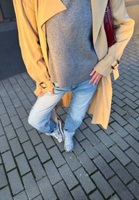 Trenci bej, pulover gri și blugi bleu deschis. Adidași argintii pe o pavele cu model. Trenciul are reveruri late și mâneci legate.