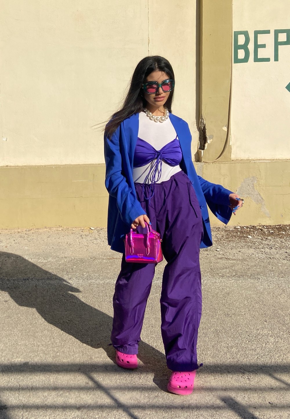 Blazer azul brilhante sobre uma blusa branca, combinado com um top curto roxo e calças largas roxas. Os acessórios incluem um colar de pérolas e uma pequena bolsa cor-de-rosa. Footwear apresenta tamancos cor-de-rosa.