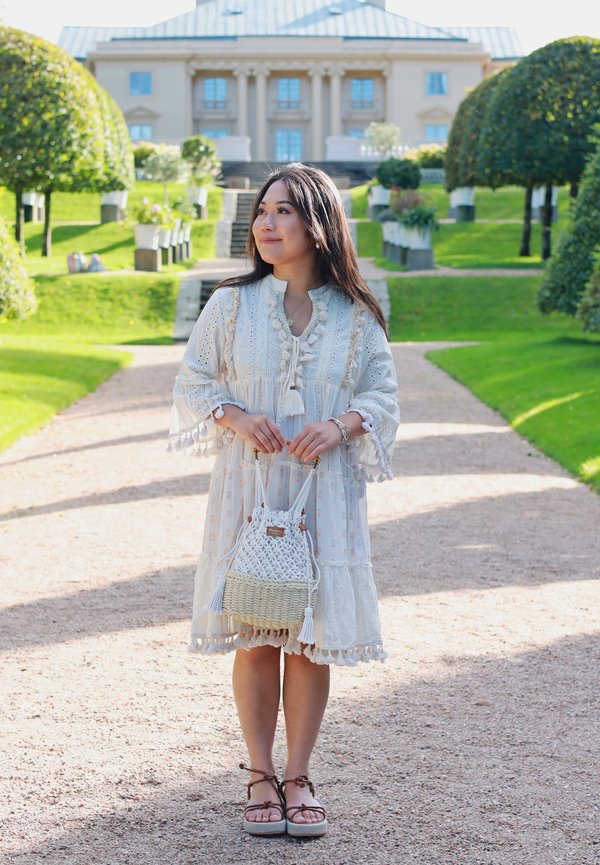 Weißes besticktes Kleid mit Quasten, kombiniert mit einer geflochtenen Handtasche mit Quastenakzent. Kiesweg mit gepflegtem Grün im Hintergrund.