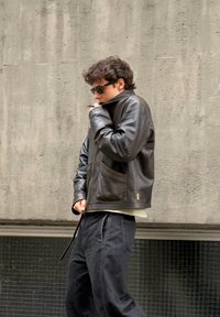 Chaqueta de cuero negra con cuello alto, múltiples bolsillos, llevada sobre una camisa clara y pantalones oscuros y sueltos. El fondo es una pared gris con textura.