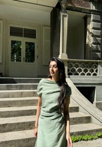 Jasnozielona lniana sukienka z falbaniastym krojem, krótkimi rękawami i luźnym fasonem. W tle widać kamienne schody i dekoracyjną architekturę.