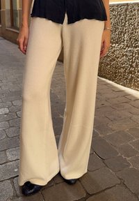 Osoba ubrana w kremowe spodnie o szerokich nogawkach, czarną bluzkę i czarne buty, stojąca na brukowanej ulicy obok teksturowanej beżowej ściany.