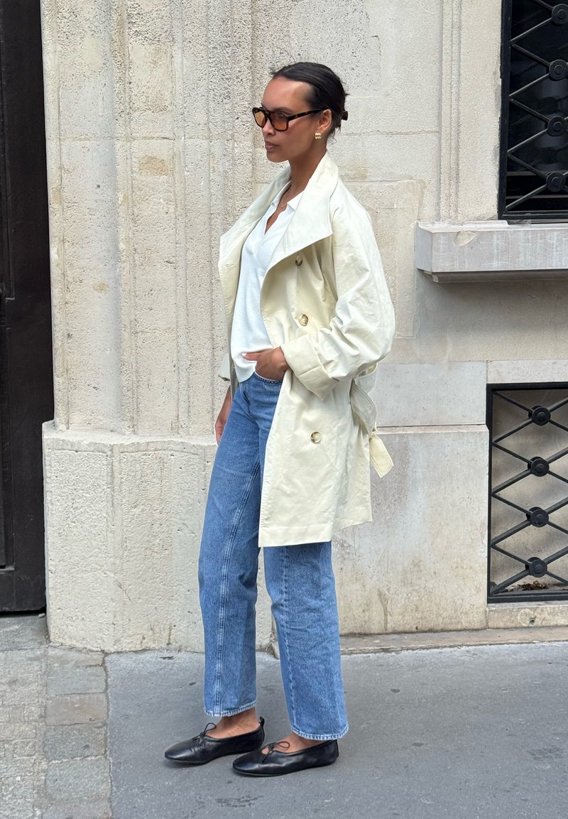 Femme aux cheveux foncés en chignon portant des lunettes de soleil, un trench crème, une chemise blanche, un jean bleu et des ballerines noires, debout sur un trottoir en ville.